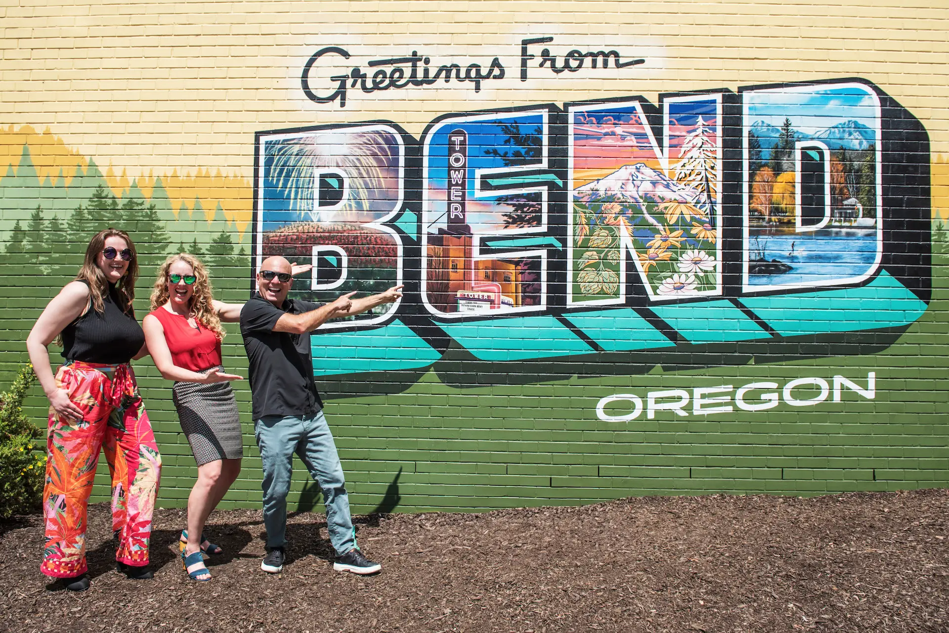 Three people posing in front of a colorful ‘Greetings from Bend, Oregon’ mural